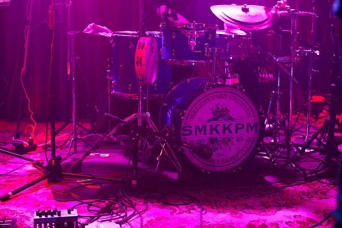 Zestaw perkusyjny podświetlony fioletowym światłem, z widocznym bębnem basowym z napisem 'SMKKPM' i datą 2023, tamburyn zawieszony na bębnie, mikrofony i kable na podłodze z wzorzystym dywanem.