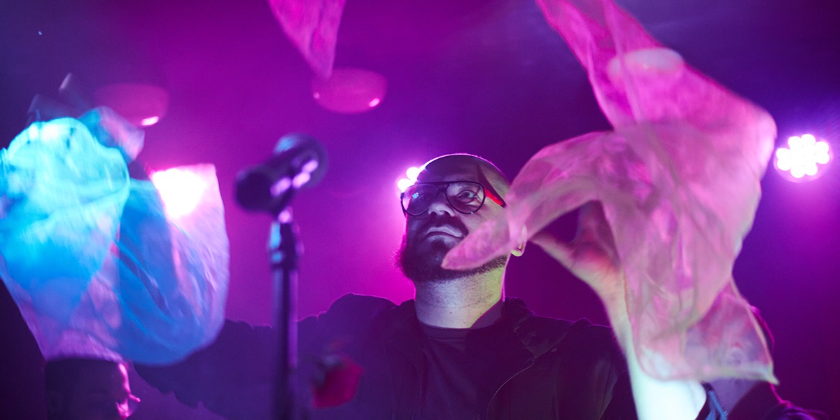 Mężczyzna z brodą i okularach, oświetlony fioletowym światłem, gestykuluje dłońmi otoczonymi przez przezroczyste materiały, mikrofon widoczny w tle.