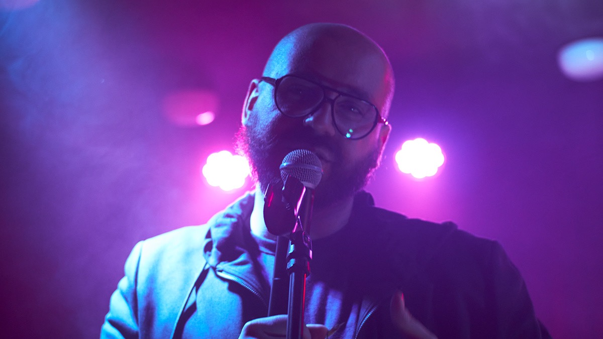 Mężczyzna z brodą i okularami śpiewa do mikrofonu na scenie w fioletowym oświetleniu.