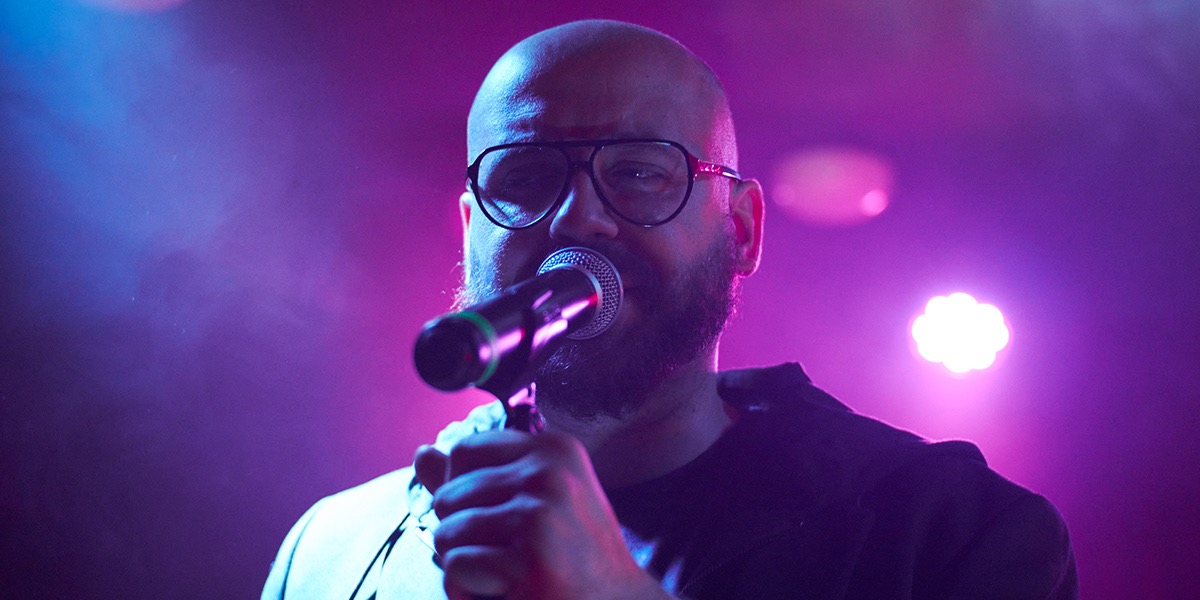 Łysy mężczyzna w okularach śpiewa do mikrofonu na scenie oświetlonej fioletowym i niebieskim światłem.