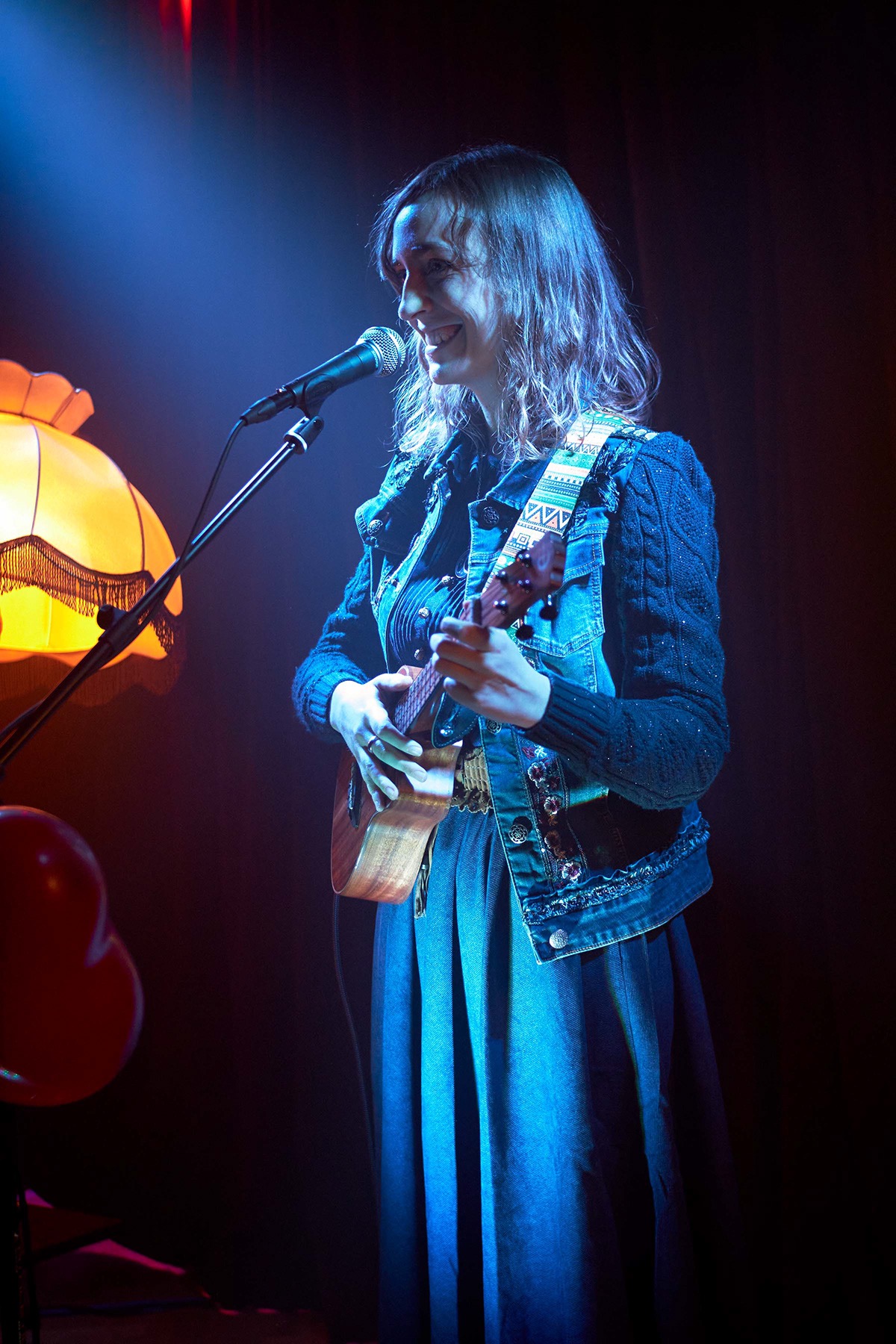 Uśmiechnięta kobieta z ukulele śpiewa do mikrofonu na scenie, oświetlona niebieskim światłem reflektora, obok lampy z frędzlami i czerwonych balonów.