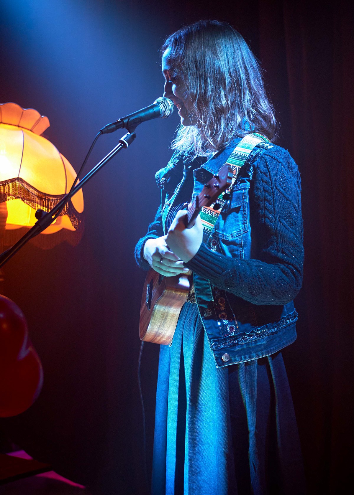 Kobieta z blond włosami śpiewa do mikrofonu grając na ukulele w oświetleniu scenicznym, obok stoi lampa z frędzlami.