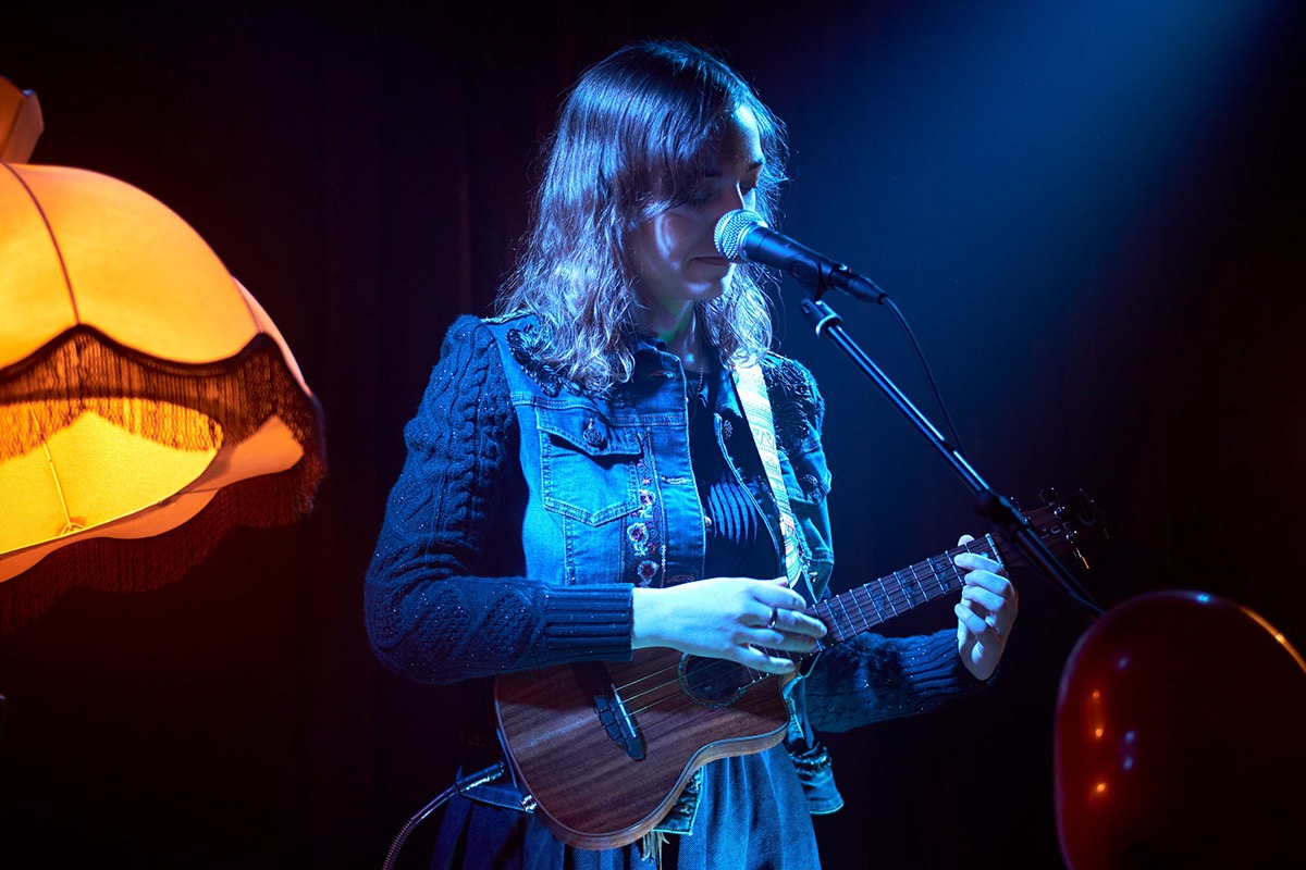 Młoda kobieta w dżinsowej kurtce gra na ukulele przy mikrofonie, oświetlona niebieskim światłem scenicznym, z żółtą lampą z frędzlami w tle.
