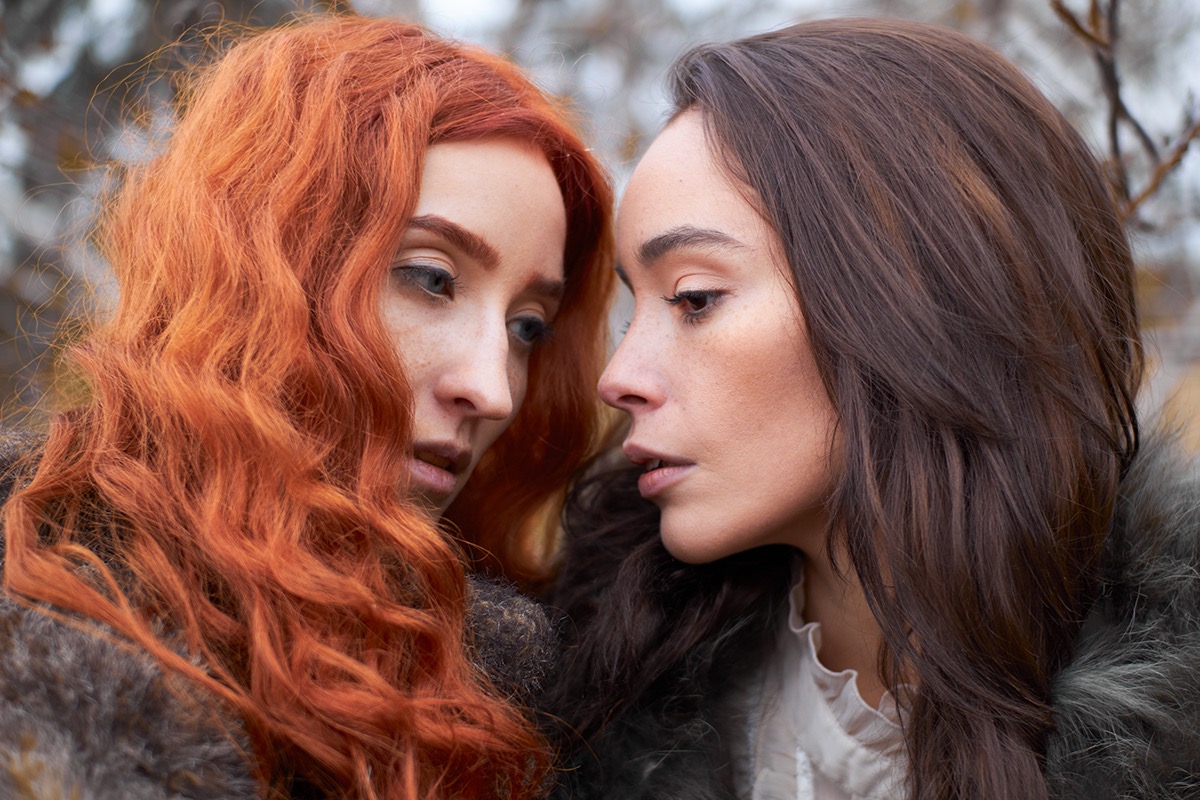 Portret dwóch kobiet z profilu, jedna z rudymi kręconymi włosami, druga z ciemnymi prostymi włosami, obie ubrane w futrzane okrycia.