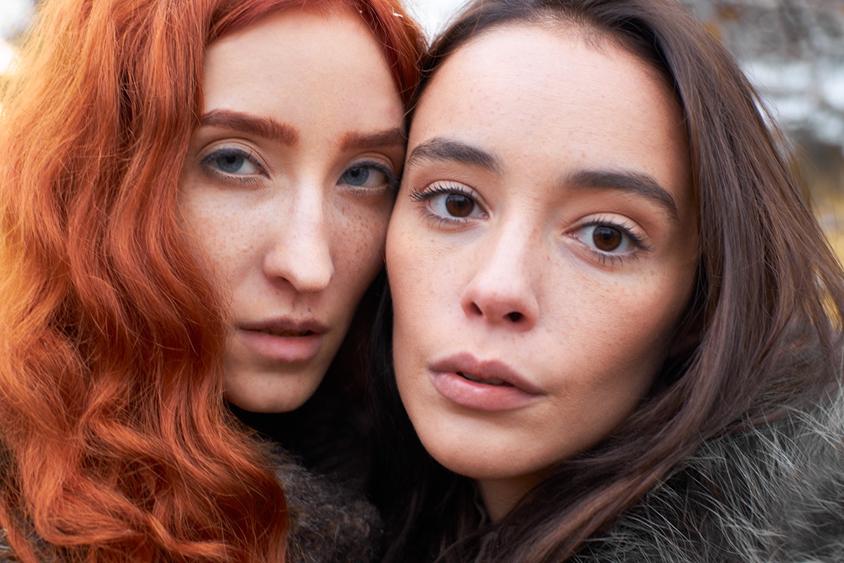 Portret dwóch kobiet z bliska: jedna z rudymi, kręconymi włosami i niebieskimi oczami, druga z ciemnymi włosami i brązowymi oczami, obie z delikatnymi piegami.