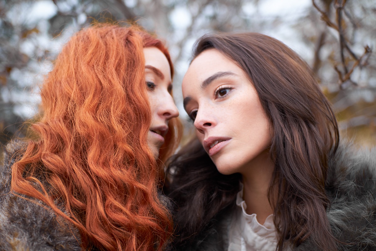 Portret dwóch młodych kobiet, rudowłosej i brunetki, w futrzanych okryciach, z delikatnym makijażem, na rozmytym tle drzew
