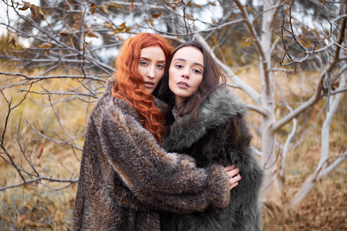 Dwie kobiety w futrzanych płaszczach, obejmujące się na tle drzew bez liści, jesienna sceneria, naturalne światło.