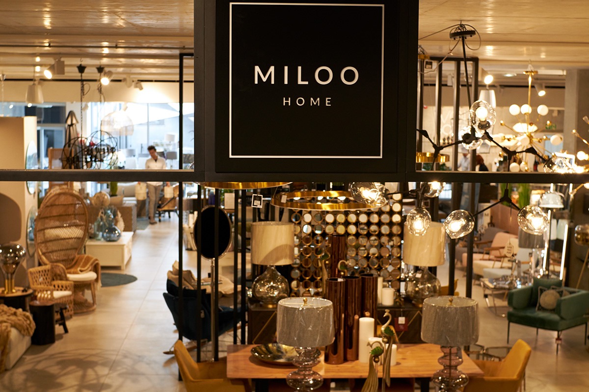 Wnętrze sklepu z wyposażeniem wnętrz, widoczne lampy, krzesła, dekoracje i logo 'MILOO HOME' na czarnym szyldzie.