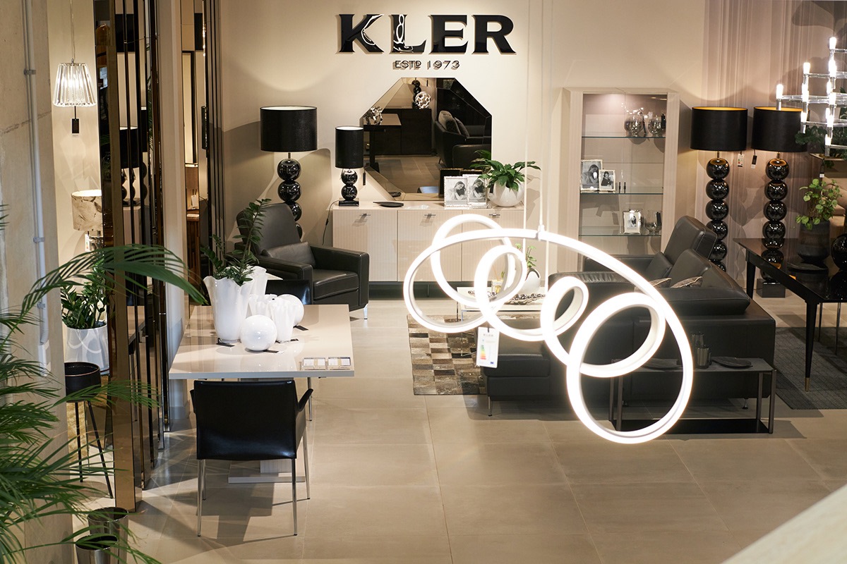 Salon meblowy Kler: czarny skórzany narożnik, stół z krzesłami, dekoracyjne lampy kuliste i modernistyczna lampa wisząca w kształcie spirali, lustro nad komodą, rośliny doniczkowe.