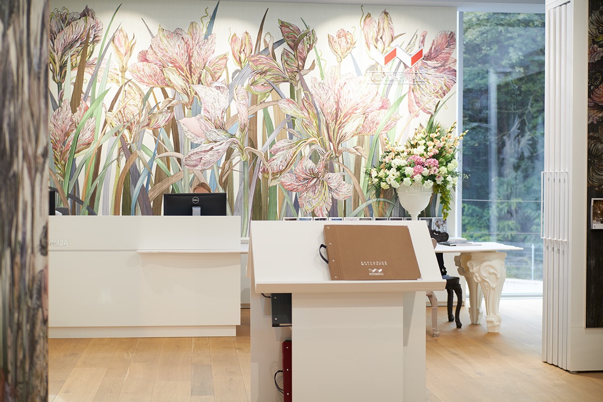 Wnętrze salonu z recepcją i ekspozycją tapet z motywem kwiatowym, widoczne biurko z komputerem, katalog tapet oraz dekoracja kwiatowa w wazonie.