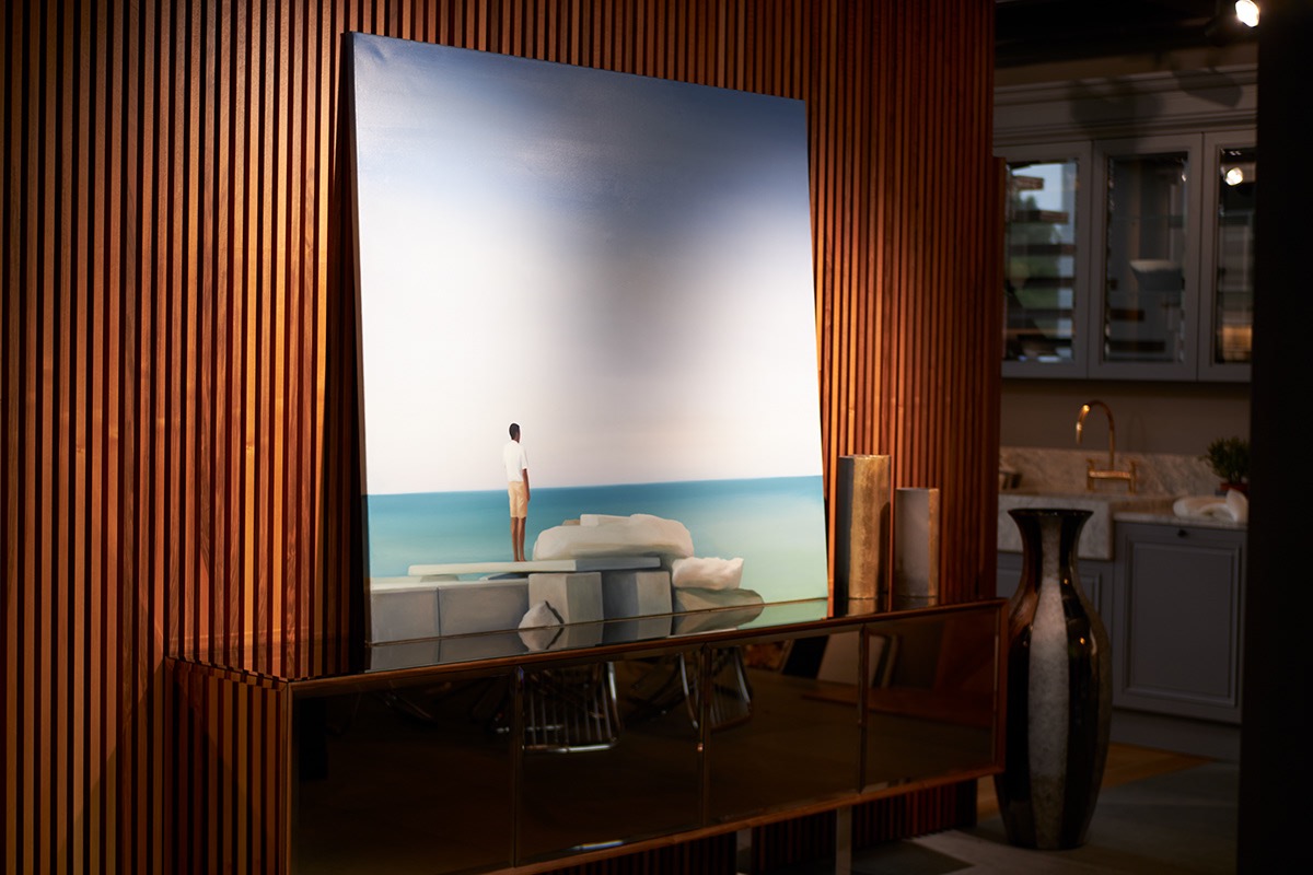 Obraz z motywem nadmorskim, przedstawiający sylwetkę mężczyzny na tle morza i nieba, umieszczony na komodzie z lustrzanym frontem w pomieszczeniu z drewnianą ścianą i widoczną kuchnią w tle.