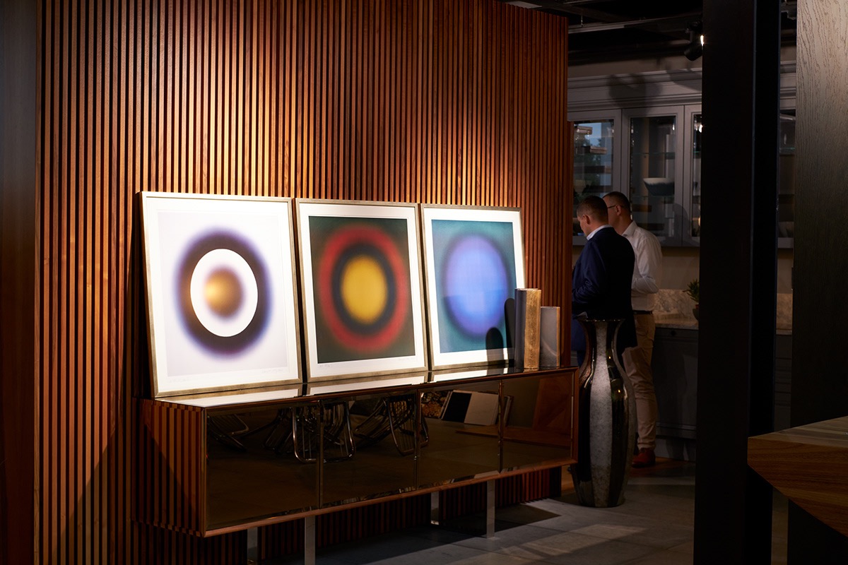 Wnętrze salonu z trzema abstrakcyjnymi obrazami w złotych ramach, stojącymi na lustrzanej komodzie na tle ściany z drewnianych listew; w tle dwóch mężczyzn rozmawia w kuchni.