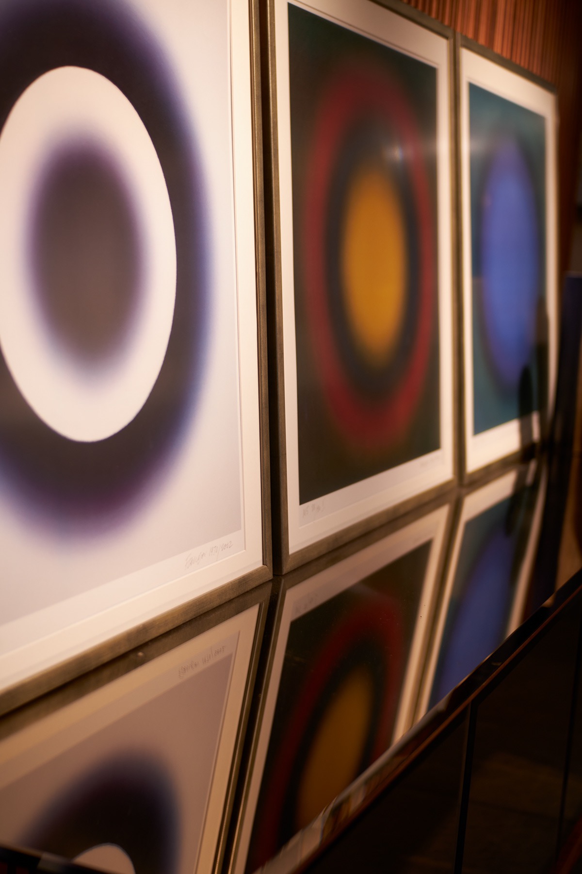 Trzy abstrakcyjne obrazy w ramkach, przedstawiające koncentryczne okręgi w odcieniach fioletu, czerwieni i niebieskiego, odbijające się w błyszczącej powierzchni poniżej.