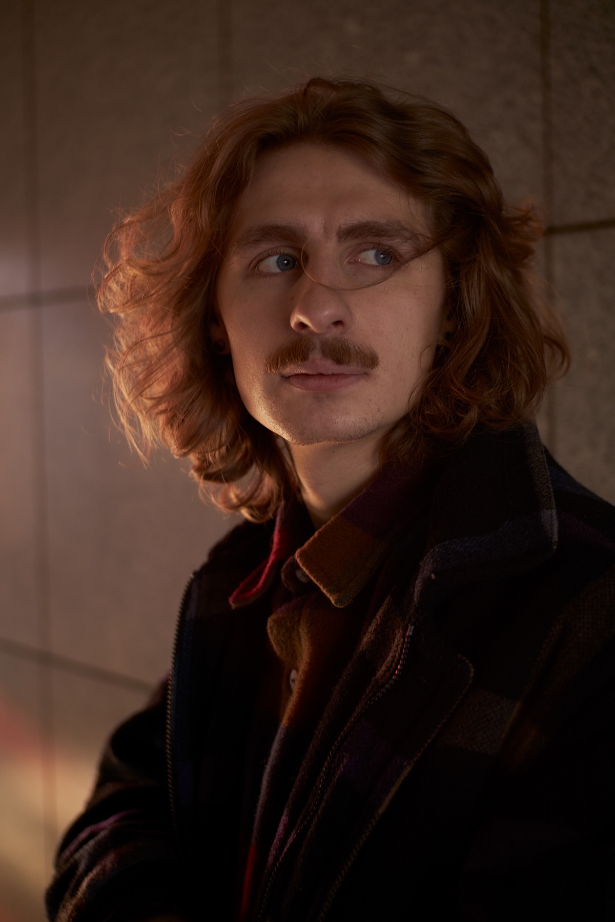 Portret młodego mężczyzny z wąsami, spoglądającego w bok, w ciepłym oświetleniu.