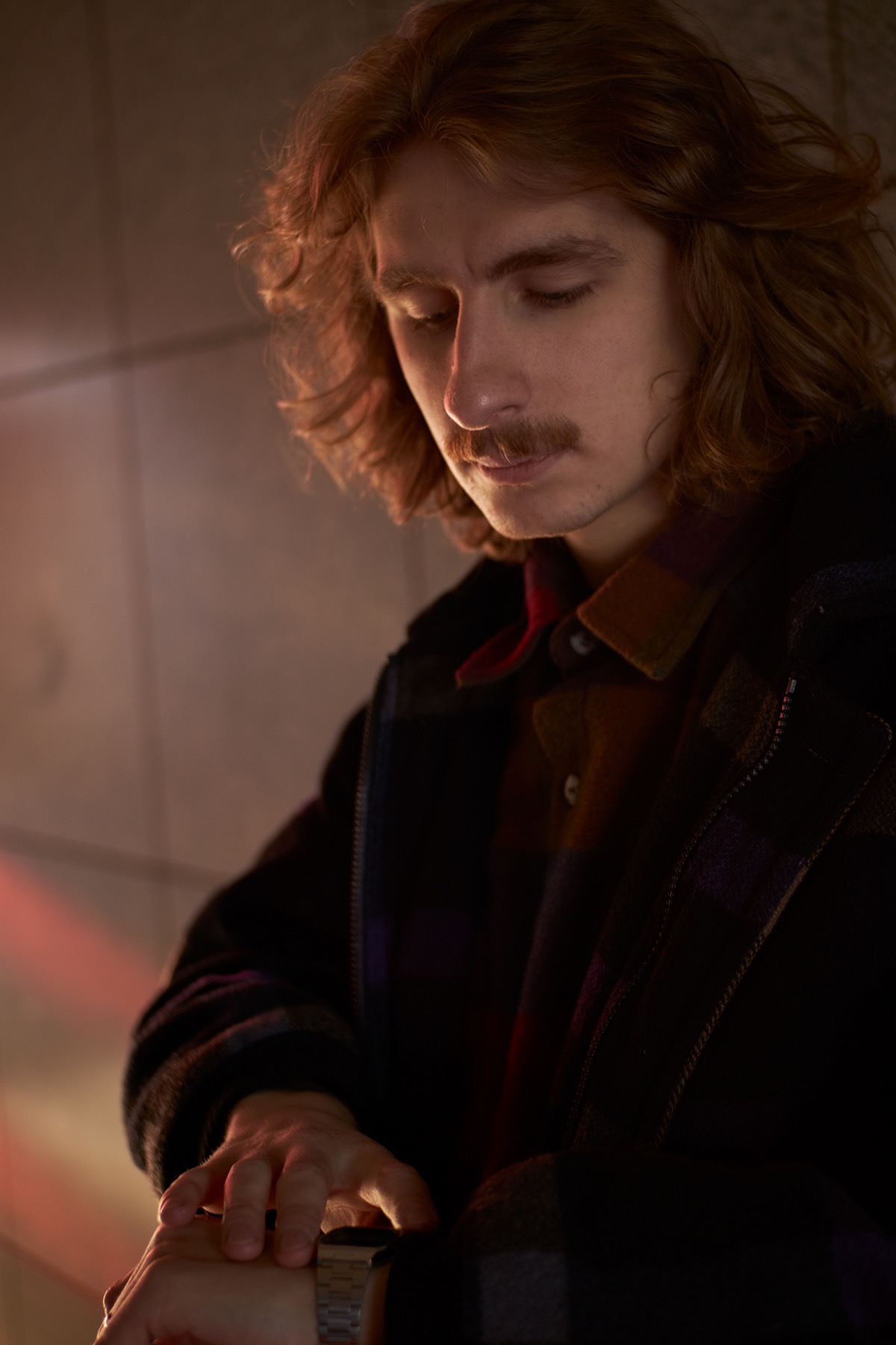 Mężczyzna z długimi włosami i wąsami spogląda na srebrny zegarek na ręce, ubrany w ciemną kurtkę i koszulę w kratę, oświetlony ciepłym światłem.