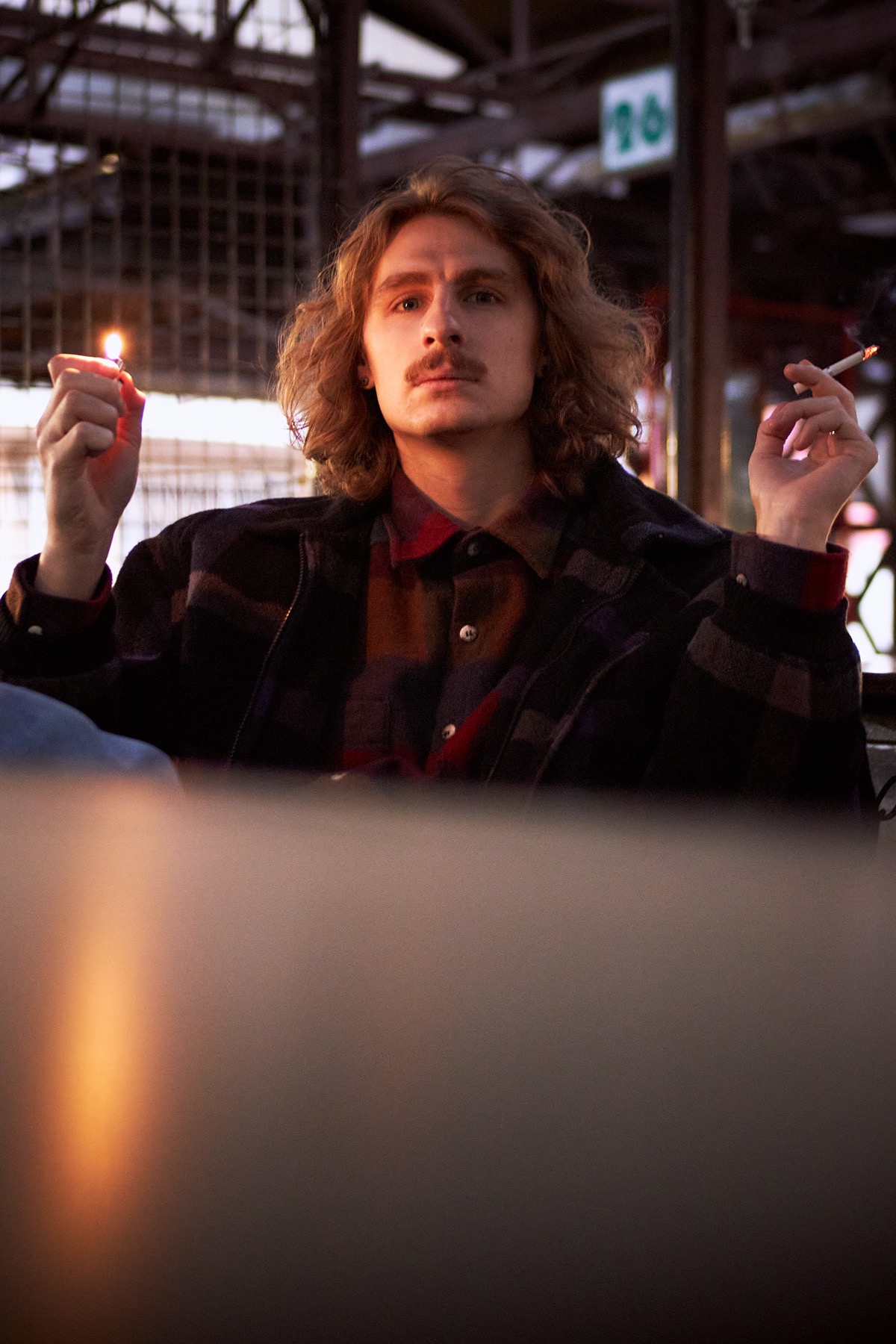 Mężczyzna z wąsami, w koszuli w kratę i kurtce, siedzi i pali papierosa, trzymając w drugiej ręce zapaloną zapałkę, w tle metalowa konstrukcja.