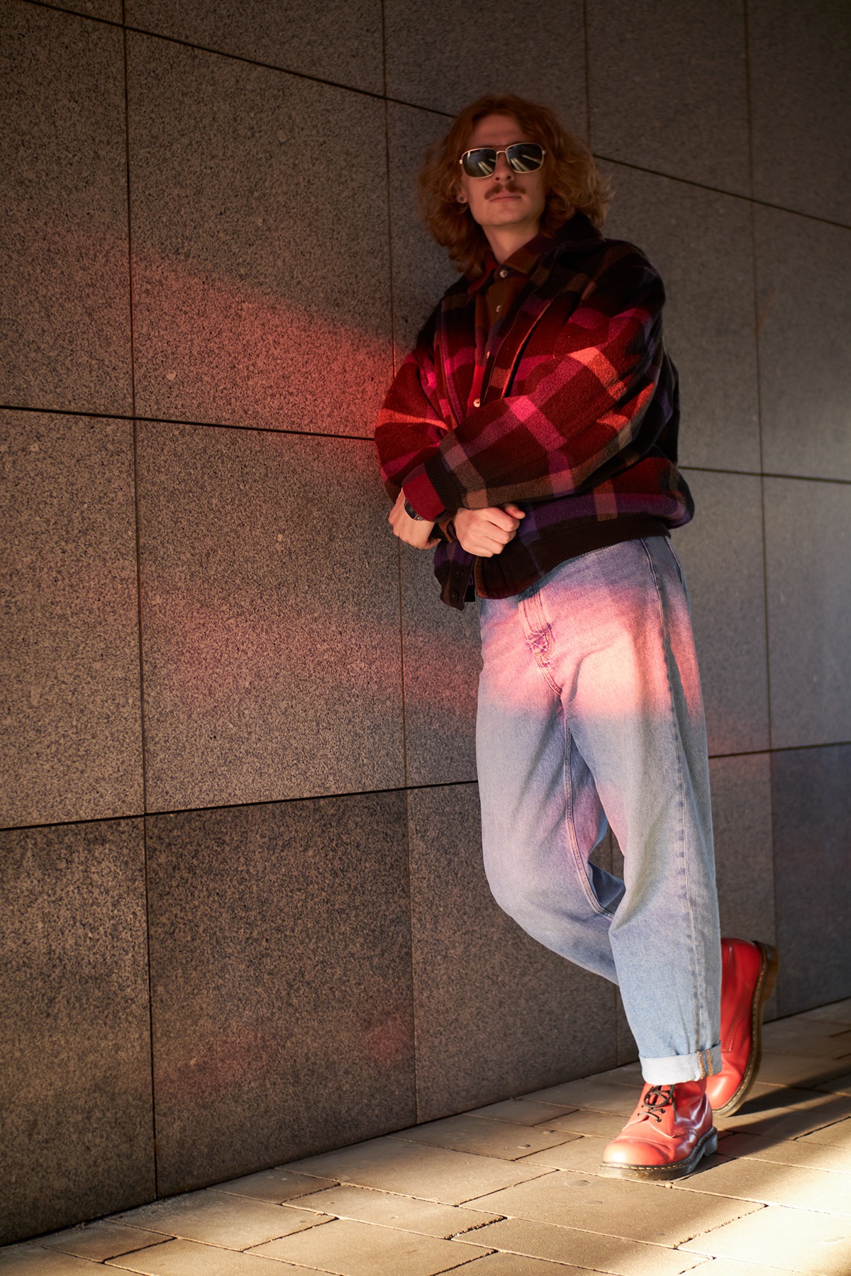 Mężczyzna w okularach przeciwsłonecznych i flanelowej kurtce opiera się o szarą ścianę z czerwoną poświatą, ubrany w dżinsy i czerwone buty.