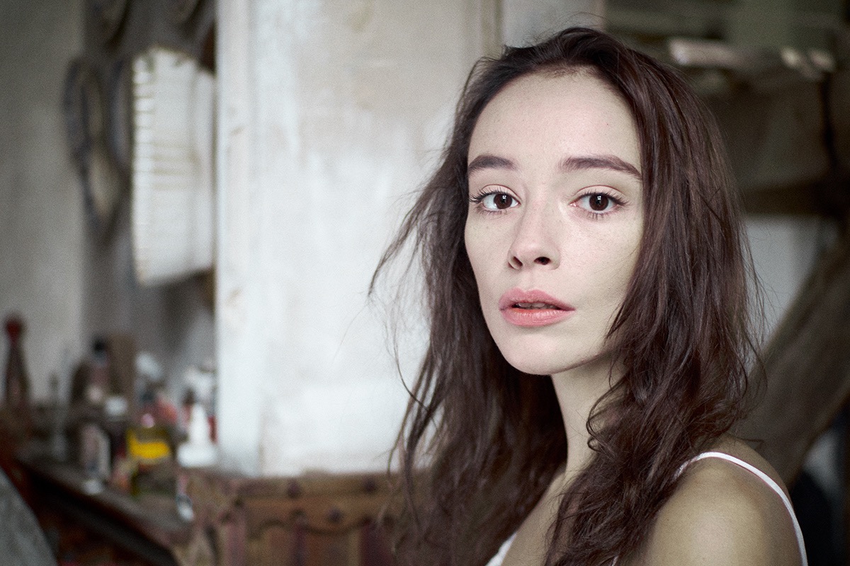 Portret młodej kobiety z rozwianymi włosami, naturalny makijaż, delikatne światło, w tle rozmyte przedmioty na półce.