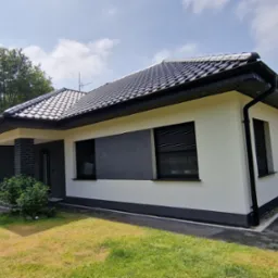 Nowoczesny dom jednorodzinny z białą elewacją, elementami szarego tynku strukturalnego i czarną dachówką, widok z boku, z zaparkowanym busem firmy budowlanej M-BUD oferującej docieplenia elewacji...