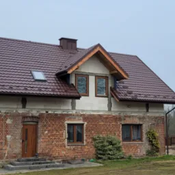Dom z nowym, brązowym dachem z blachodachówki i oknem dachowym. Częściowo ocieplony budynek z ceglaną fasadą i drewnianą stolarką okienną.