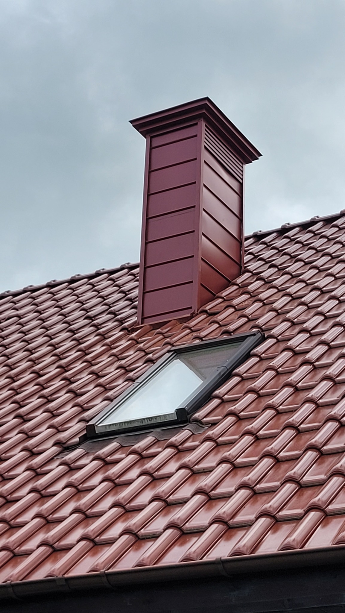 Czerwony dach z dachówki ceramicznej z oknem połaciowym i kominem obłożonym blachą w kolorze dachówki, na tle pochmurnego nieba. Bliski kadr.