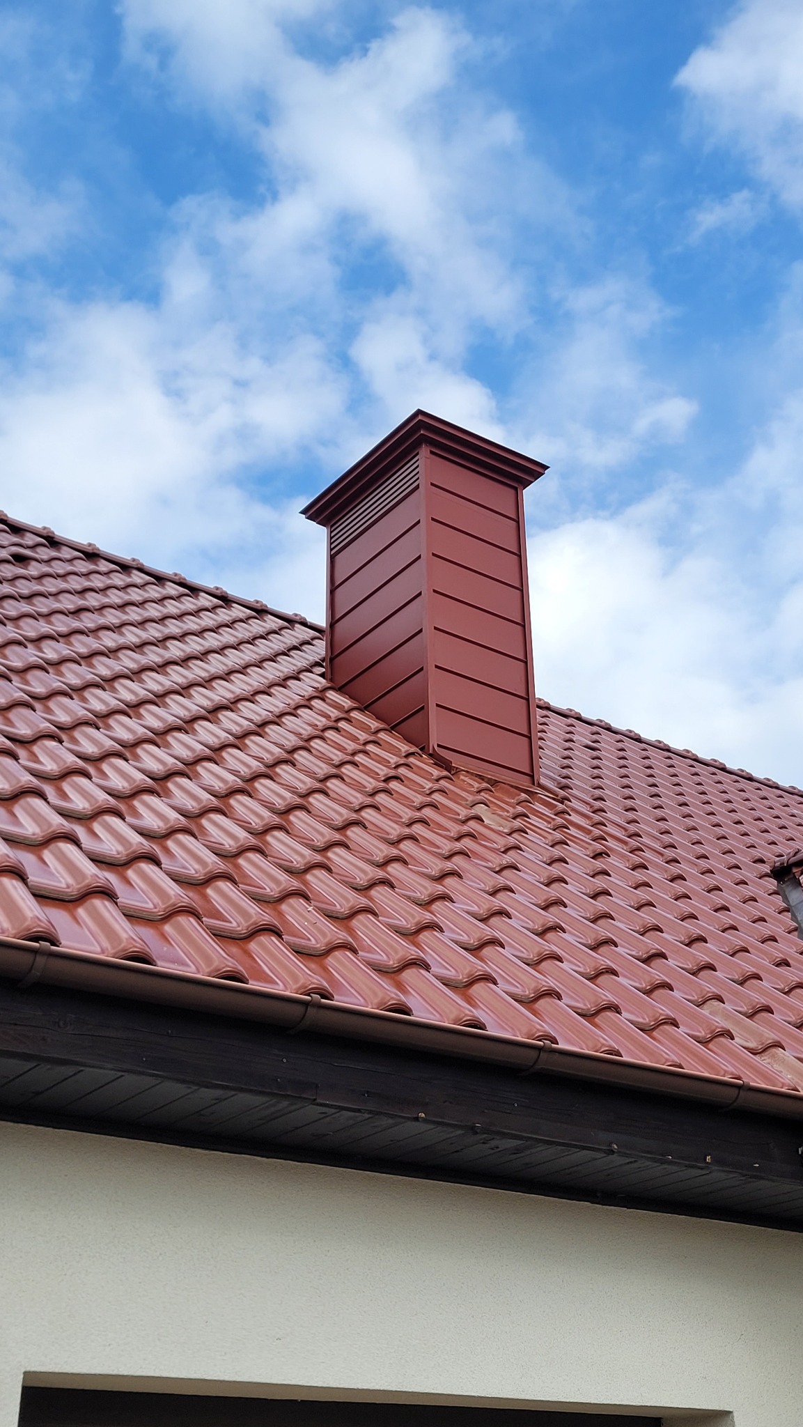 Czerwona dachówka ceramiczna z kominem wentylacyjnym na tle błękitnego nieba z chmurami. Widoczny fragment elewacji i rynna w kolorze dachu.