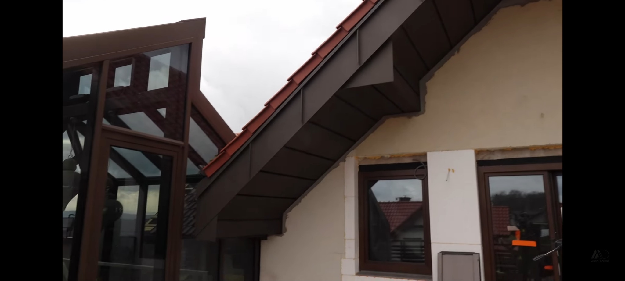 Nowe, brązowe obróbki blacharskie na dachu domu z ciemnymi oknami. Widoczne elementy wykończeniowe elewacji i konstrukcja szklanej werandy.