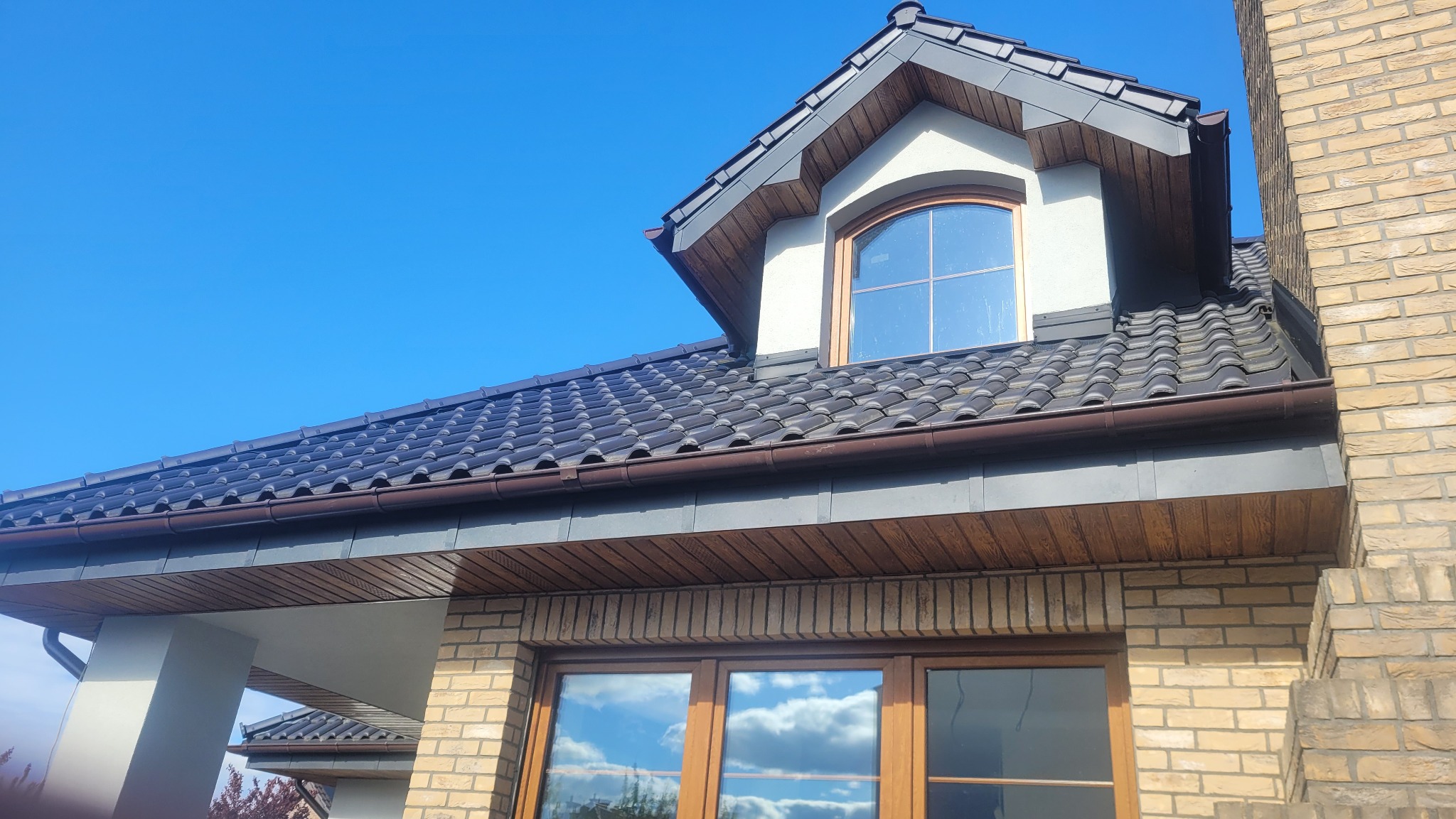 Ciemna dachówka karpiówka na dachu domu z lukarną i brązowymi rynnami, widok z dołu na tle błękitnego nieba.