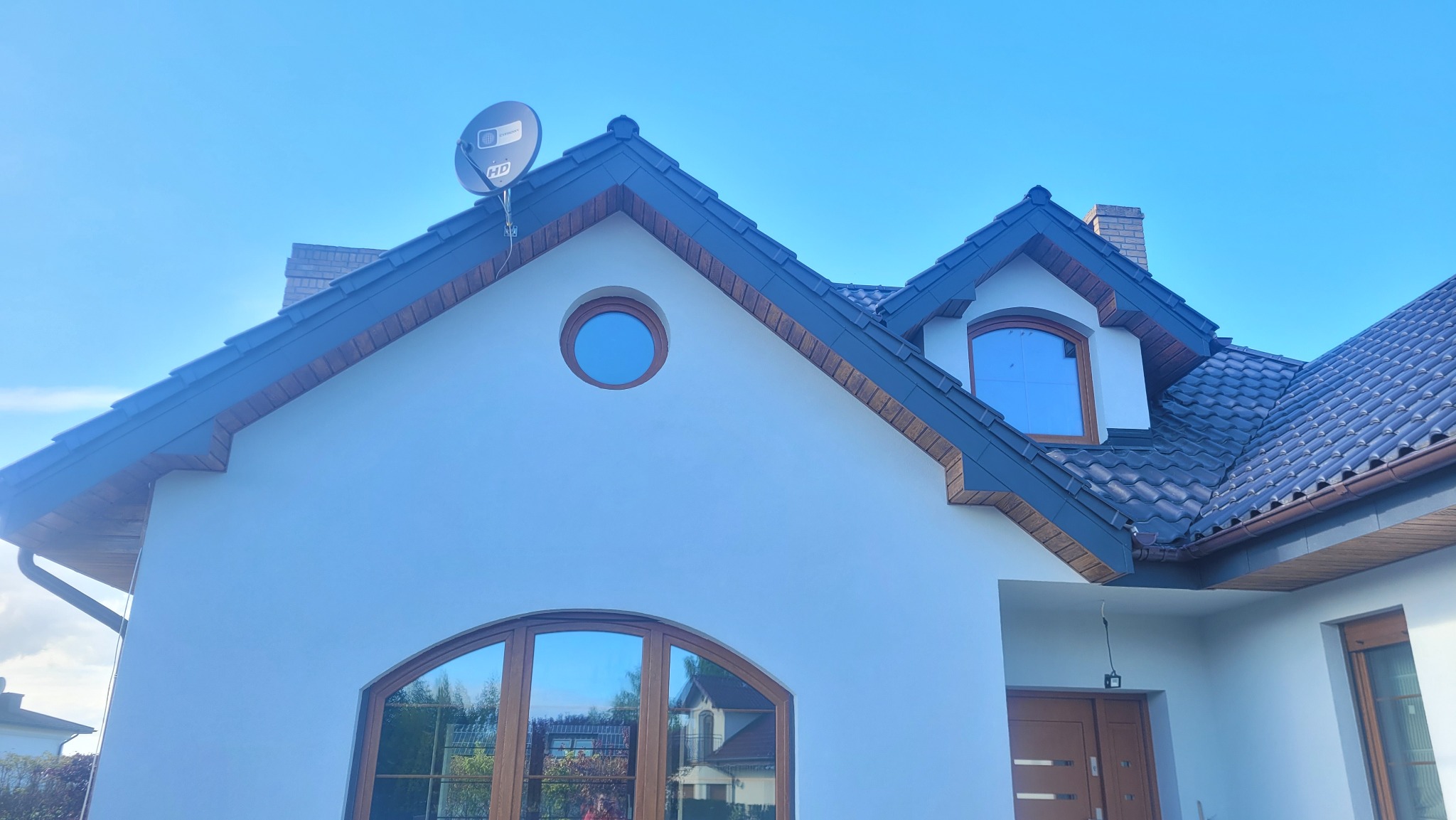 Dom jednorodzinny z ciemną dachówką, okrągłym oknem na poddaszu i satelitą na dachu. Widoczne rynny i obróbki blacharskie.