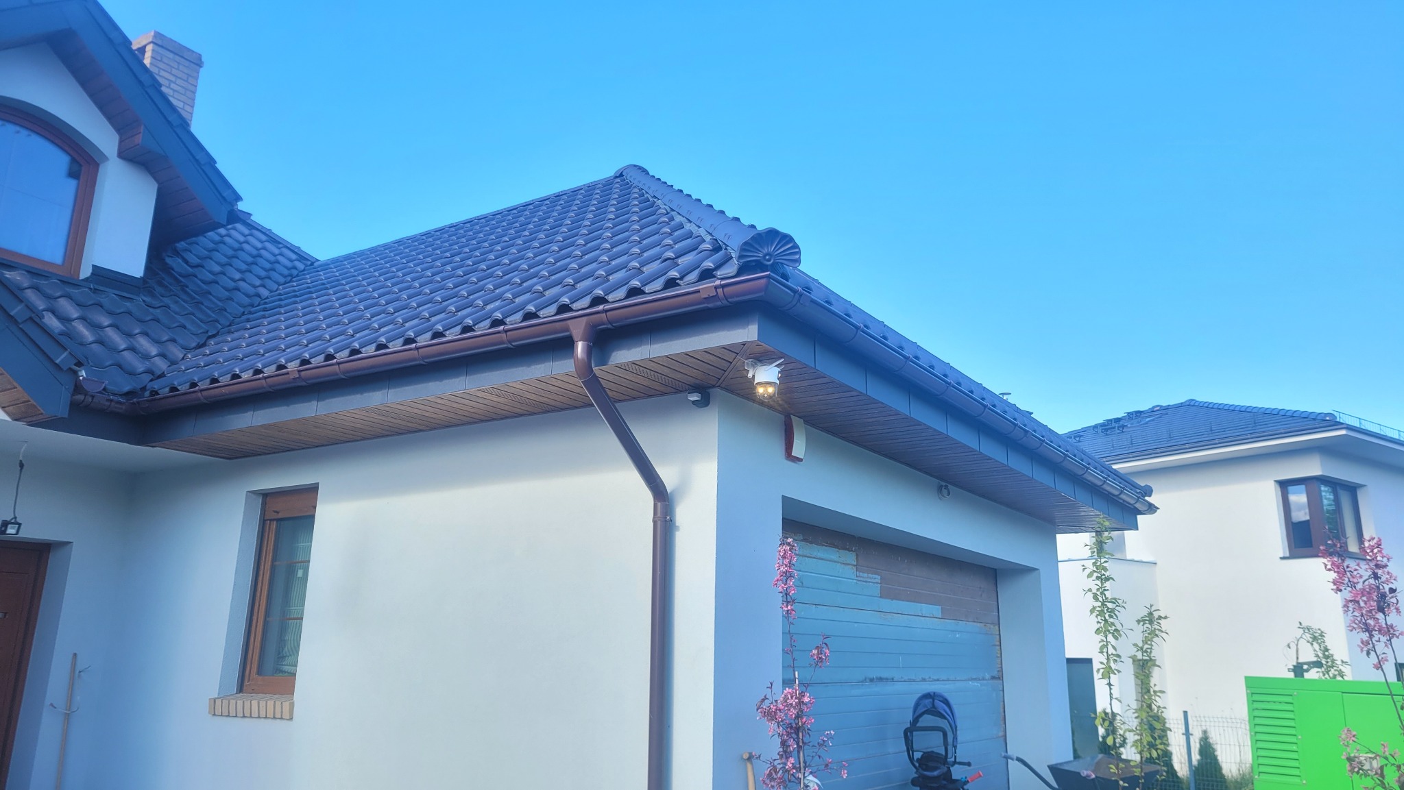 Nowoczesny dom z ciemną dachówką i brązowymi rynnami w słoneczny dzień. Widoczne detale wykończenia dachu i elewacji. Uwagę przykuwa oświetlenie zewnętrzne.