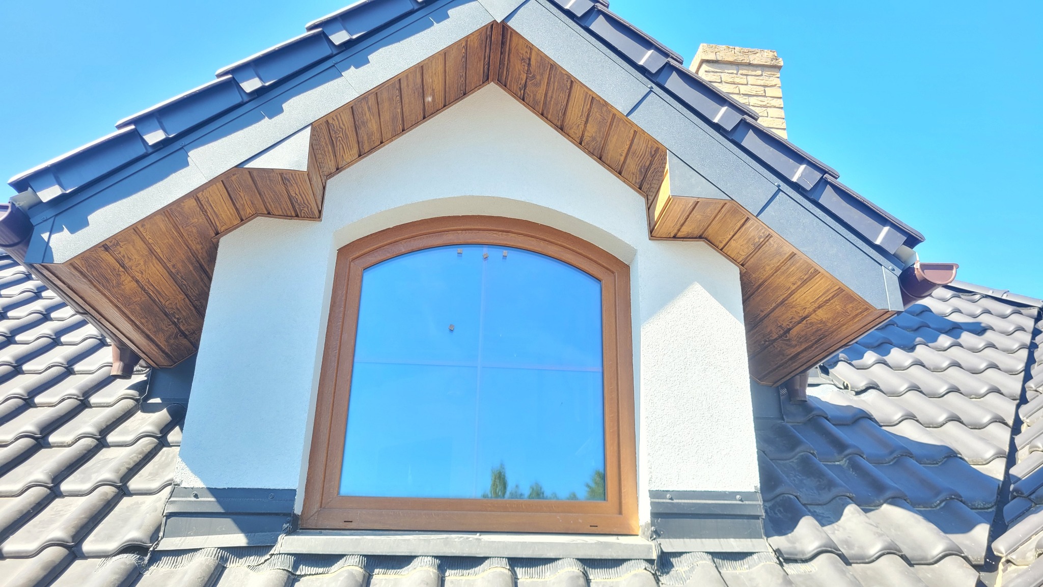 Szczegółowe ujęcie lukarny z brązową stolarką okienną i drewnianą obudową, na tle ciemnej dachówki i błękitnego nieba. Widoczny komin w tle.