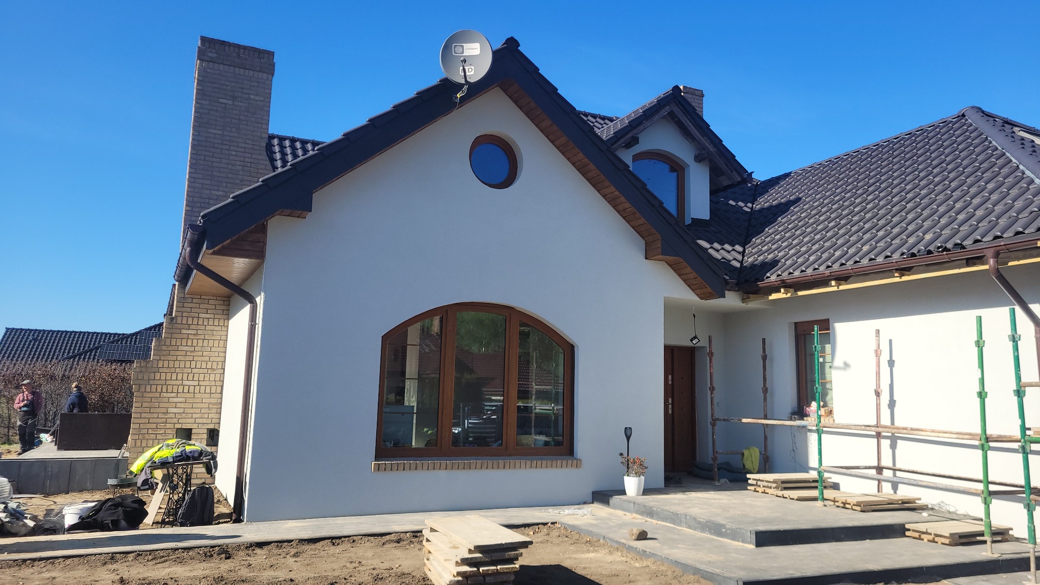 Dom jednorodzinny z ciemną dachówką i nową elewacją. Widoczna antena satelitarna na dachu, okrągłe okno i fragment rusztowania. Prace wykończeniowe.