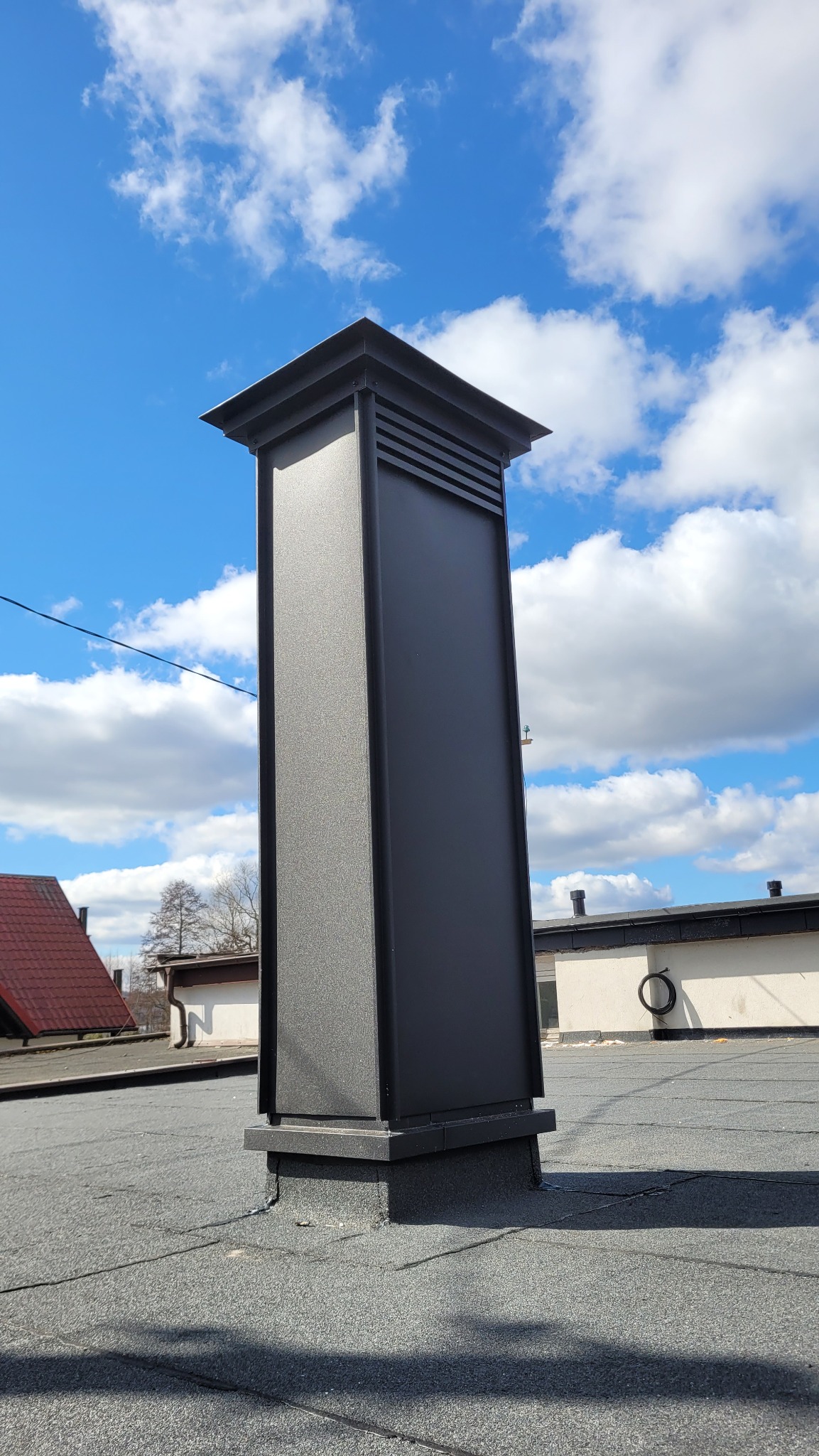 Nowoczesny, grafitowy komin wentylacyjny na dachu pokrytym papą, na tle błękitnego nieba z białymi obłokami. Widoczne fragmenty sąsiednich budynków.