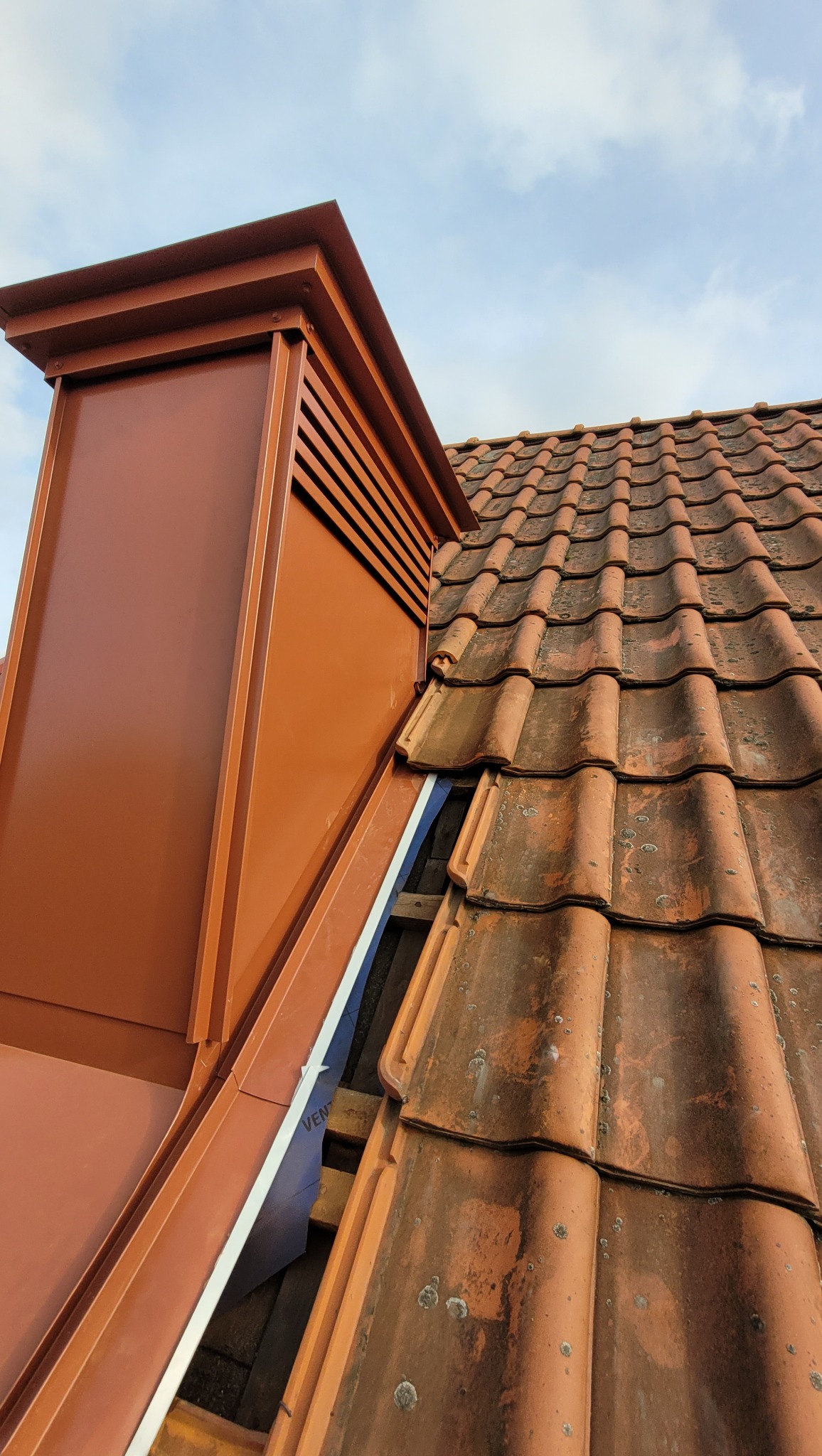 Ceglasty komin wentylacyjny z blachy trapezowej, połączony z dachem krytym starą dachówką ceramiczną, na tle błękitnego nieba.