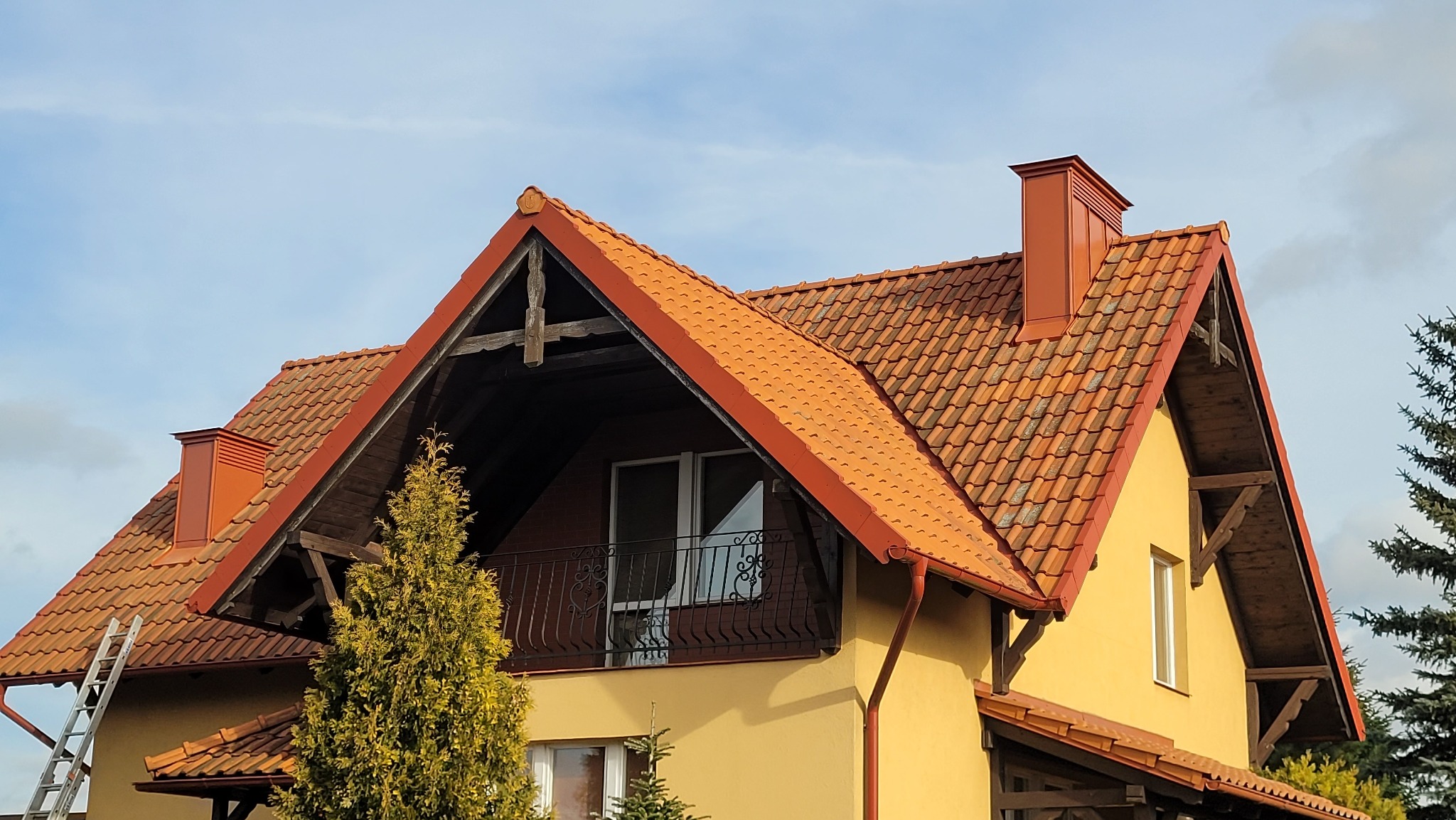 Żółty dom z czerwonym dachem z dachówki ceramicznej, balkonem i rynnami. Widoczny fragment drabiny oparty o ścianę. Ujęcie z dołu, w tle błękitne niebo.