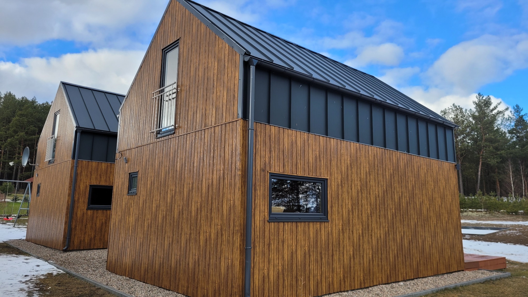 Dwa nowoczesne domy z drewnianą elewacją i ciemnymi, metalowymi dachami. Widoczne rynny i obróbki blacharskie. Tło stanowi las i fragment nieba z chmurami.