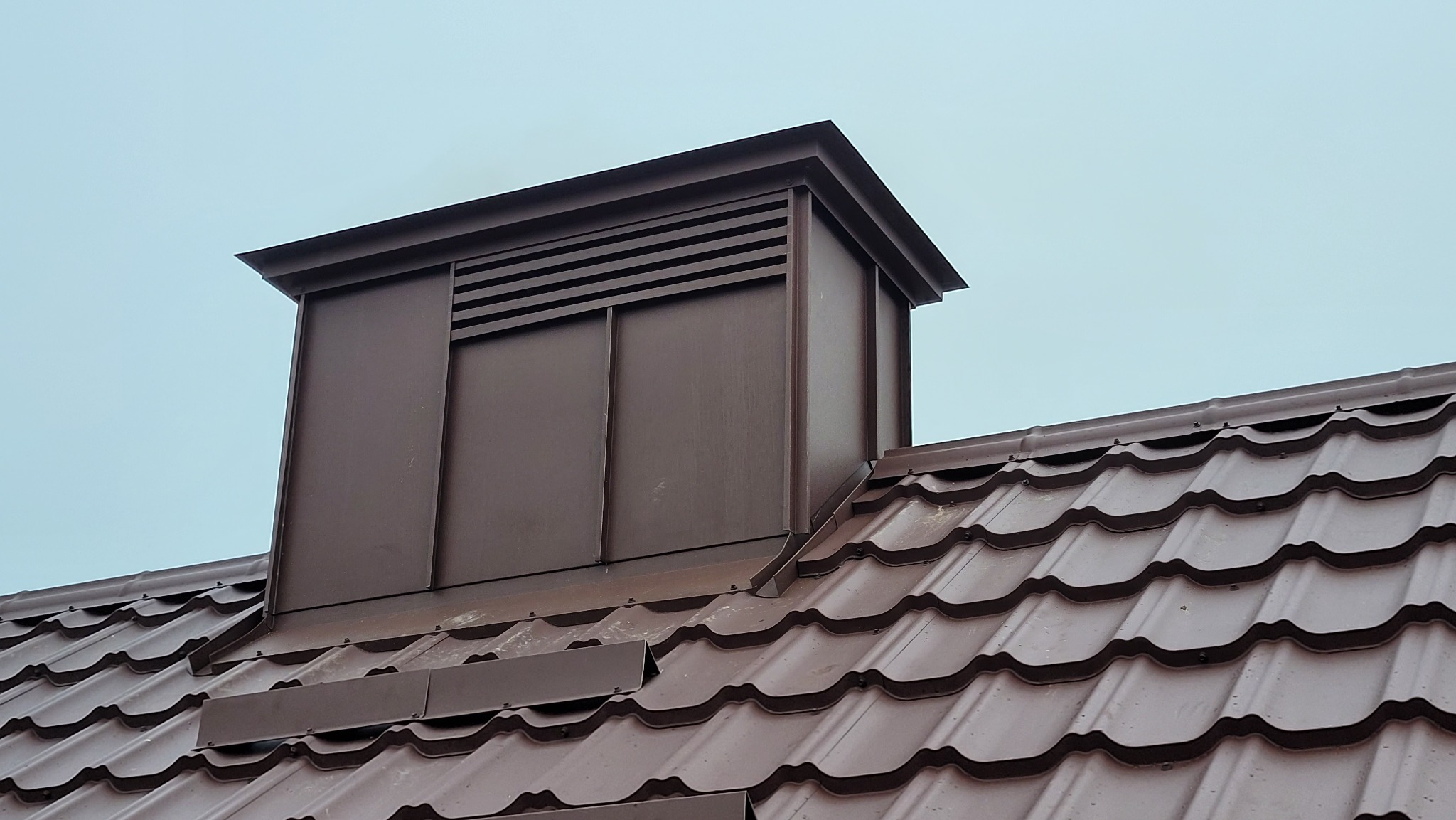 Brązowy komin wentylacyjny na dachu z blachodachówki w kolorze czekolady. Widok na górną część dachu i komin na tle pochmurnego nieba.