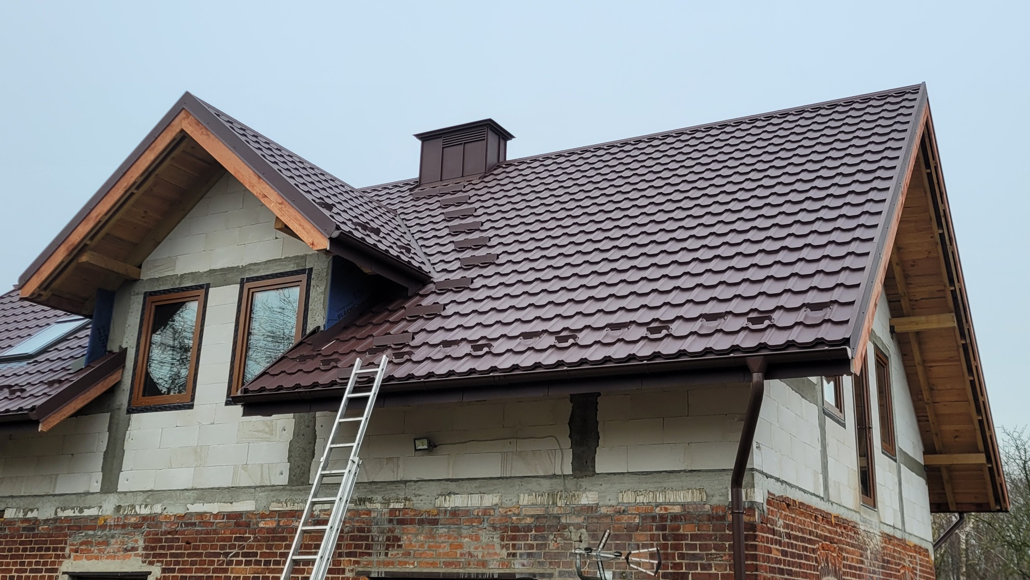 Brązowy dach z blachy trapezowej na domu w trakcie budowy, widoczna drabina oparta o ścianę, elementy drewniane więźby dachowej, ceglana podmurówka.