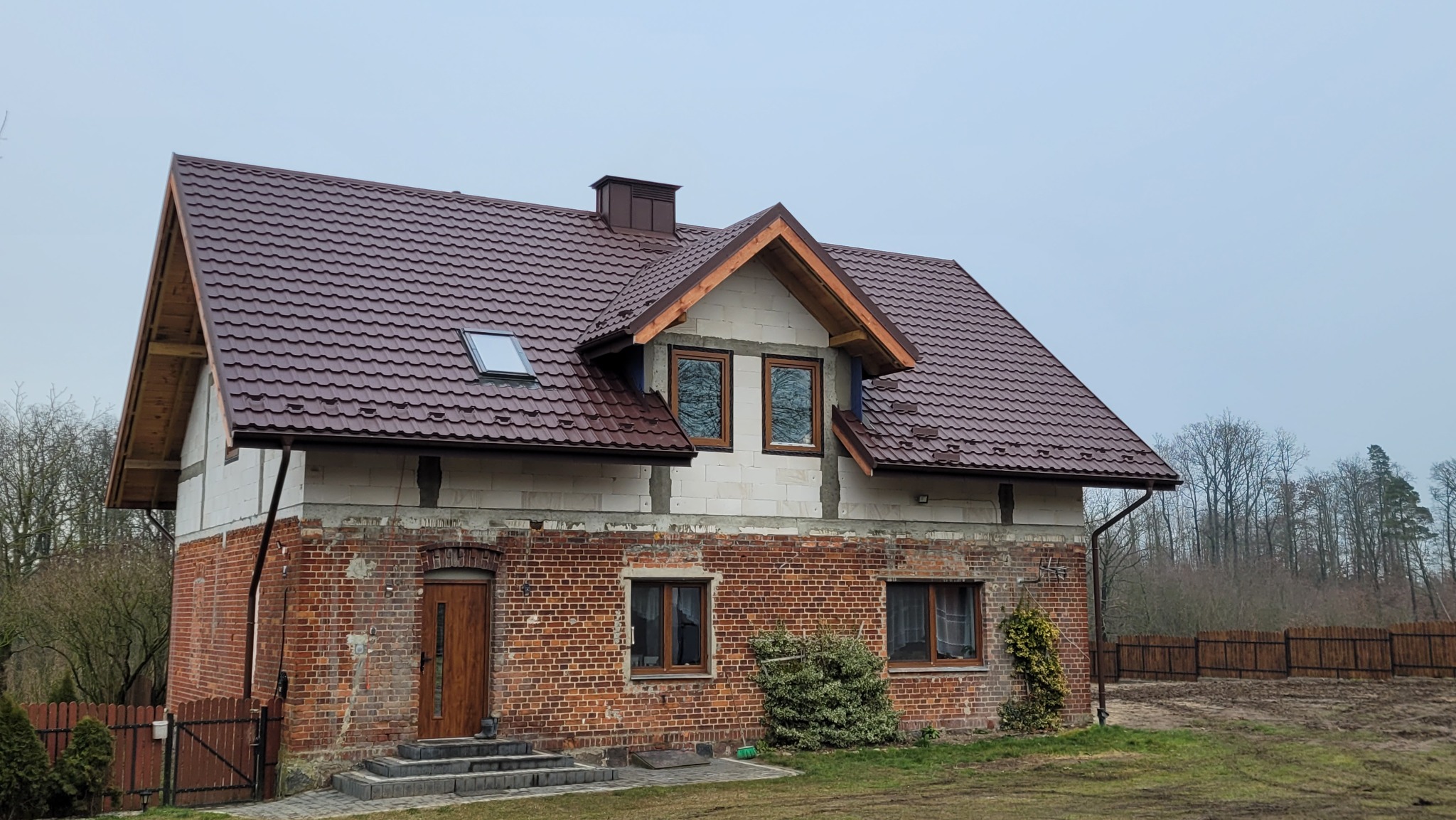 Dom z nowym, brązowym dachem z blachodachówki i oknem dachowym. Częściowo ocieplony budynek z ceglaną fasadą i drewnianą stolarką okienną.