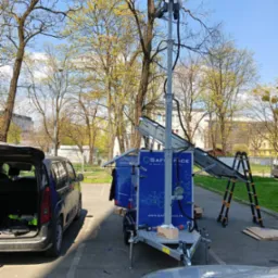 Mobilna wieża oświetleniowa z panelami solarnymi na przyczepce, zaparkowana na parkingu, z rozłożonymi panelami i drabiną obok, w tle drzewa i budynki.