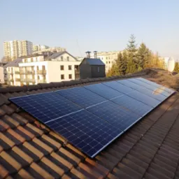 Panel słoneczny zamontowany na brązowym dachu z dachówki ceramicznej, w tle budynki mieszkalne i błękitne niebo.