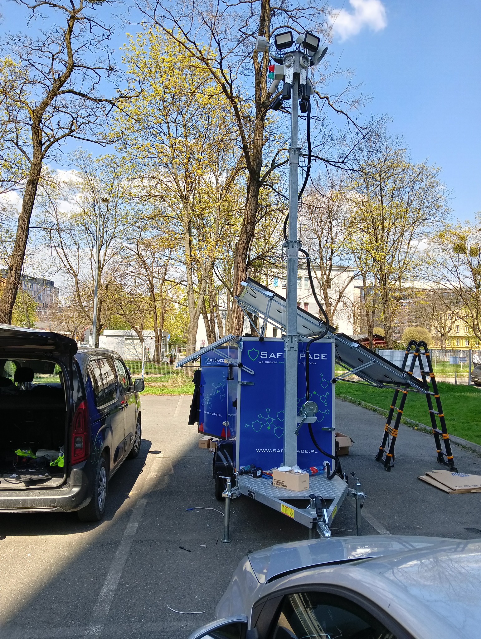 Mobilna wieża oświetleniowa z panelami solarnymi na przyczepce, zaparkowana na parkingu, z rozłożonymi panelami i drabiną obok, w tle drzewa i budynki.
