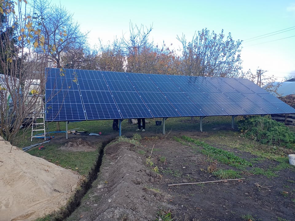 Montaż paneli fotowoltaicznych na metalowej konstrukcji w ogrodzie, widoczny wykop pod kable, drabina oparta o konstrukcję, częściowo pochmurne niebo.