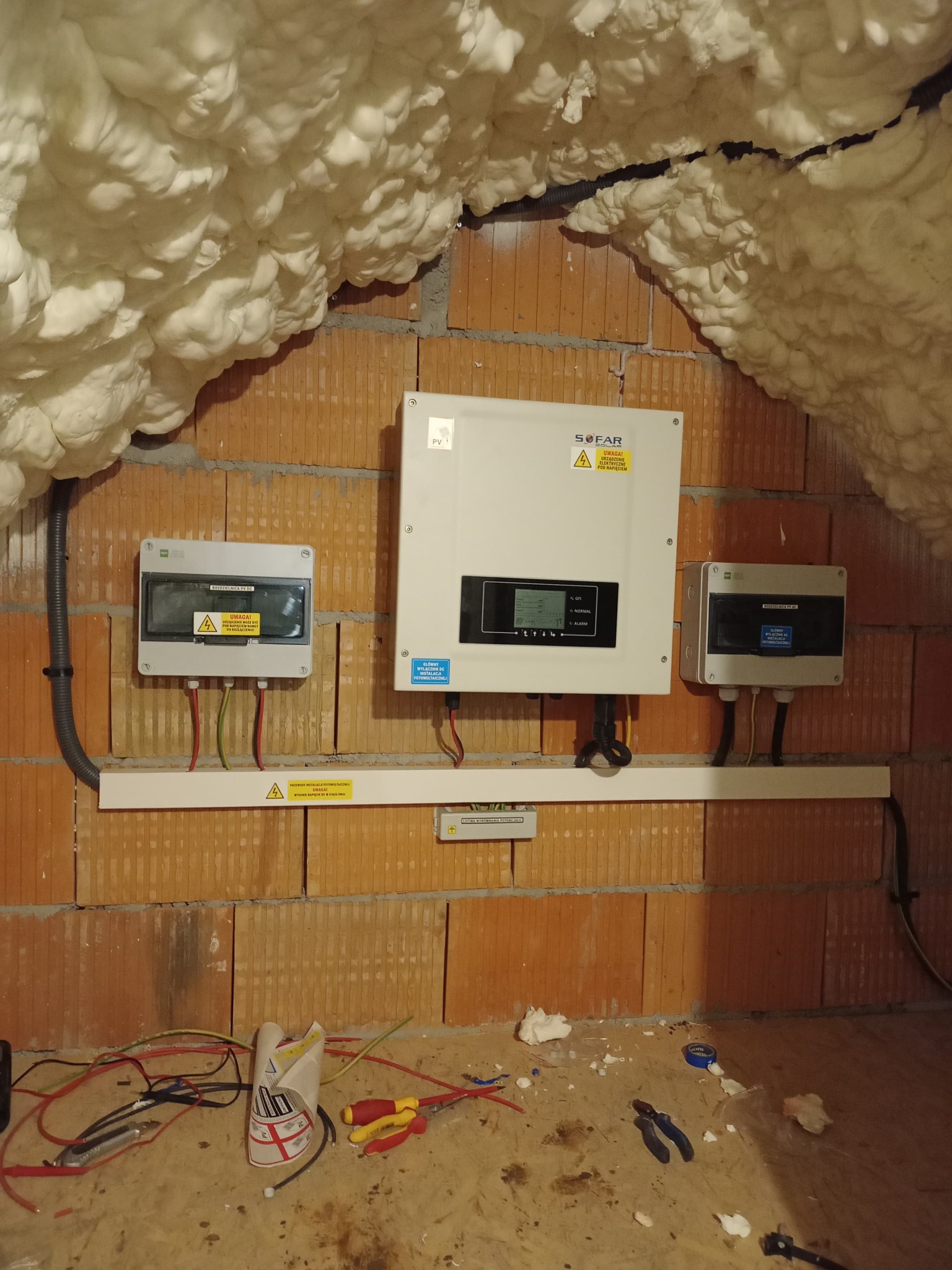 Instalacja fotowoltaiczna w trakcie montażu: widoczne rozdzielnice DC i falownik SOFAR na ceglanej ścianie, przewody i narzędzia leżące na podłodze, izolacja piankowa na suficie.