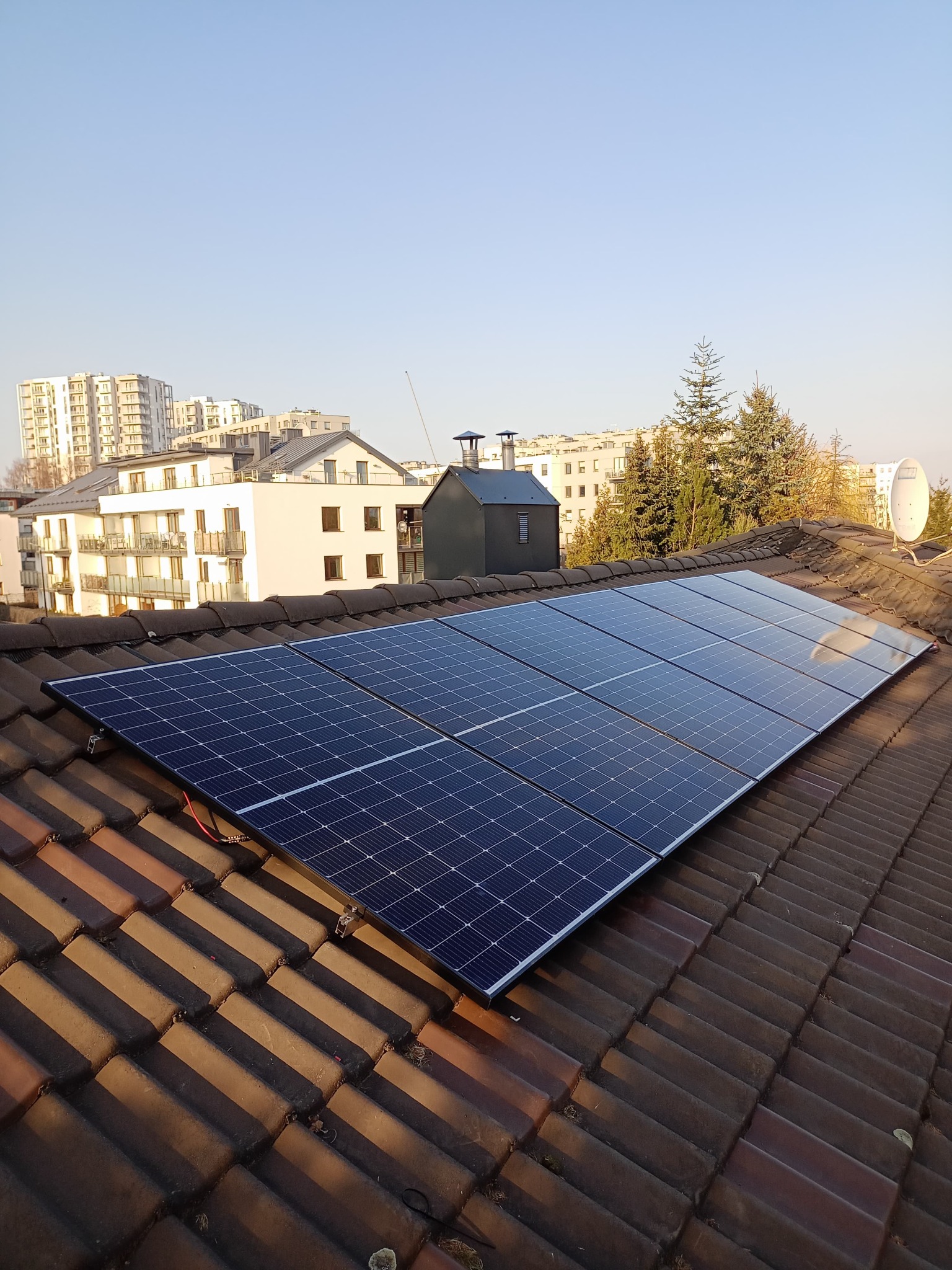 Panel słoneczny zamontowany na brązowym dachu z dachówki ceramicznej, w tle budynki mieszkalne i błękitne niebo.