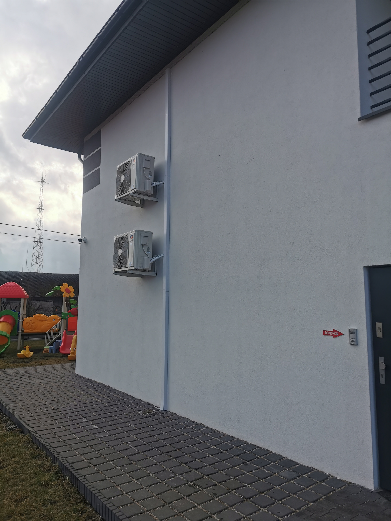 Dwa klimatyzatory zamontowane na ścianie budynku z placem zabaw w tle, widoczne z poziomu chodnika.