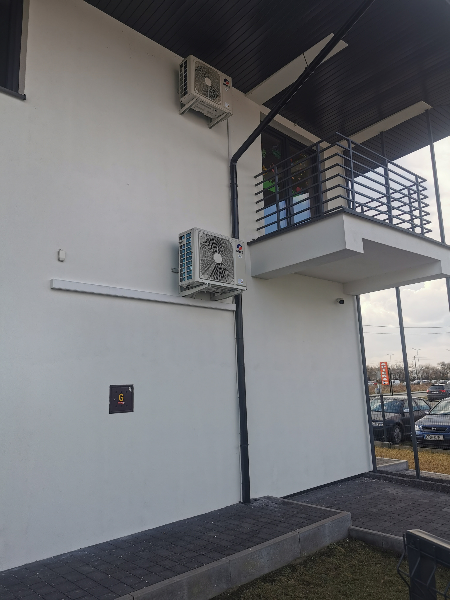 Dwa klimatyzatory Gree zamontowane na białej ścianie budynku z balkonem. Widoczna instalacja odprowadzająca skropliny. Na dole ściany skrzynka elektryczna z literą G.