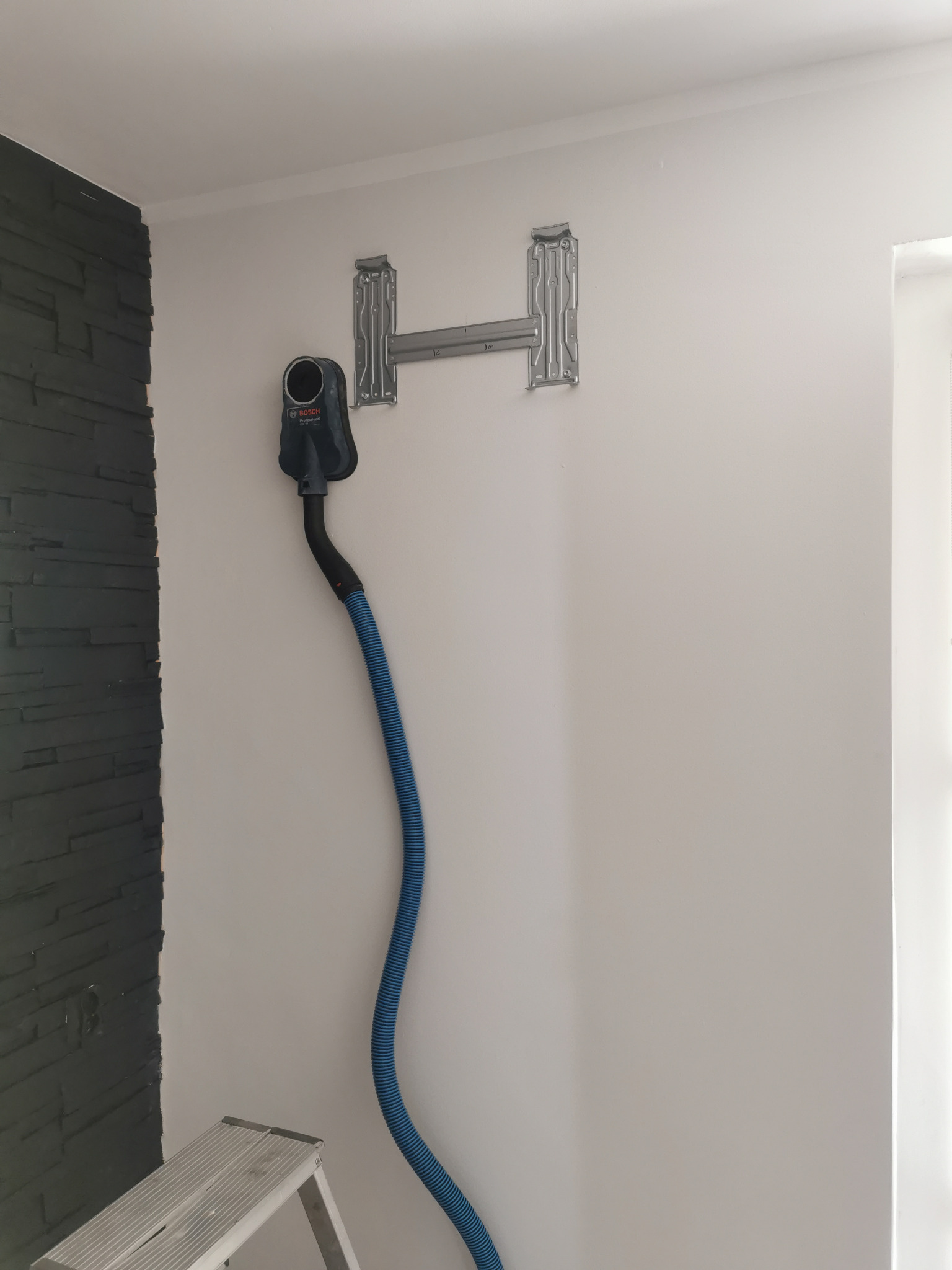 Przygotowanie do montażu jednostki wewnętrznej: metalowy wspornik na białej ścianie, podłączony odkurzacz Bosch z niebieskim wężem do wiercenia bezpyłowego.