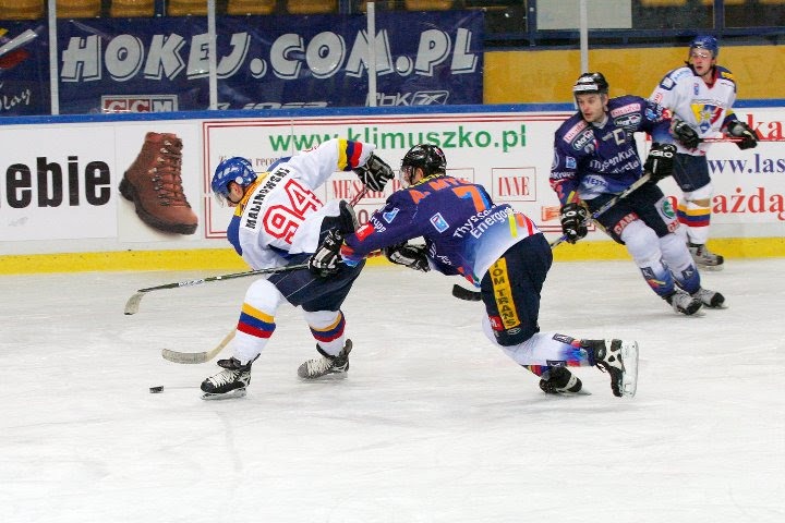 Dynamiczna scena z meczu hokeja na lodzie: zawodnik w białym stroju z numerem 94 walczy o krążek z zawodnikiem w granatowym stroju, w tle widoczni inni gracze i banery reklamowe.