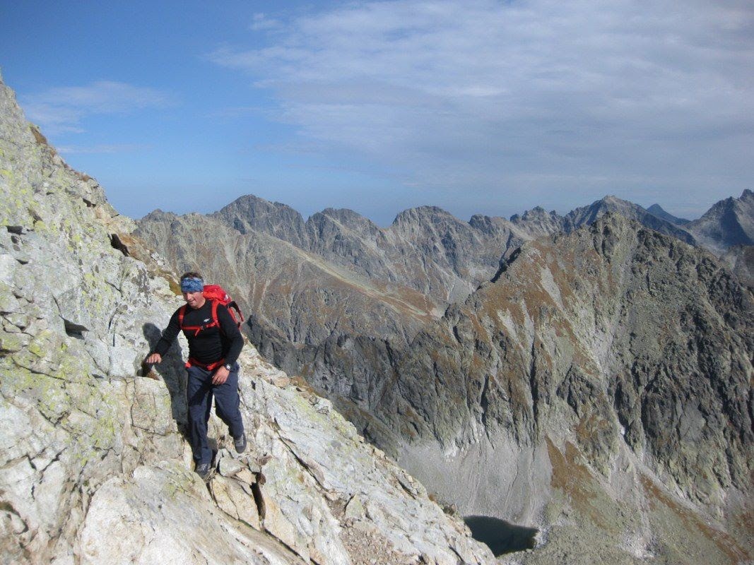 Mężczyzna z czerwonym plecakiem wędruje po skalistym zboczu góry, w tle widoczne szczyty górskie i błękitne niebo z chmurami.